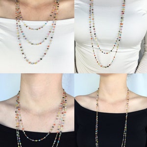 May include: A multi-strand necklace with small, colorful beads. The necklace is layered and features a variety of colors, including red, orange, yellow, green, blue, purple, and pink. The necklace is shown on a model wearing a white shirt and a black shirt.