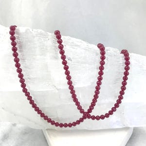 May include: A strand of small, round, faceted ruby-colored beads arranged in a draped pattern. The beads have a consistent deep red hue and a reflective surface, creating a uniform appearance along the entire length of the stringing material.