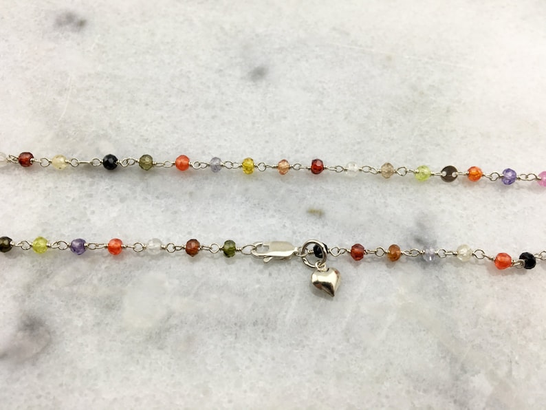 May include: A silver chain necklace with a heart charm. The chain is adorned with small, colorful beads in shades of red, orange, yellow, green, blue, and purple.