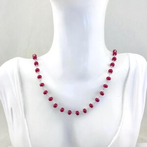 May include: A necklace of small, round, bright red beads strung on a silver chain. The necklace is displayed on a white mannequin bust, highlighting its simple elegance and vibrant colour. The beads are evenly spaced.