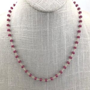 Può includere: Una delicata collana con piccole perline rotonde rosso scuro infilate su una catena d'argento. Le perline sono distanziate in modo uniforme, creando un design semplice ma elegante. La collana è esposta su un manichino.