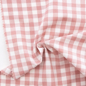 May include: Close-up of a fabric with a classic gingham pattern in white and dusty rose. The fabric is folded, showing the texture and weave. The edges are raw, and the fabric is likely cotton or linen.