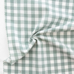 May include: A piece of sage green and white gingham fabric with a raw edge. The fabric is folded, showing the texture and pattern of the checkered design. The fabric is suitable for sewing projects.