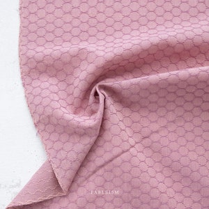 May include: Close-up of a dusty rose fabric with a textured, honeycomb-like pattern. The fabric is gathered in soft folds, revealing the texture and color variations. The word "FABLEISM" is printed in white on the lower right corner.