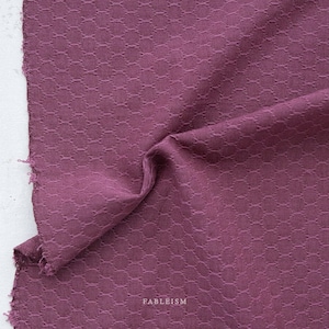 May include: Close-up of a piece of fabric in a deep plum color. The fabric has a textured, geometric pattern and is slightly crumpled, with a raw edge visible on the left side. The word "FABLEISM" is printed in white at the bottom.