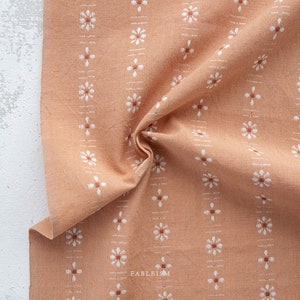 May include: Peach-colored fabric with a repeating pattern of white floral and brown cross designs. The fabric is folded, showing texture and detail. The word "FABLEISM" is printed in the lower right corner.