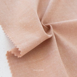 May include: Close-up of a swatch of peach-colored fabric with a textured weave. The fabric is folded and draped, revealing the texture and color variations. The edge of the fabric is cut with a zig-zag pattern. The word "FABLEISM" is printed on the bottom right.