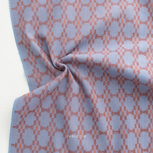May include: A close-up of a folded fabric with a repeating geometric pattern. The fabric is a light blue color with a pattern of orange squares and rectangles. The word "FABLEISM" is printed in white on the fabric.