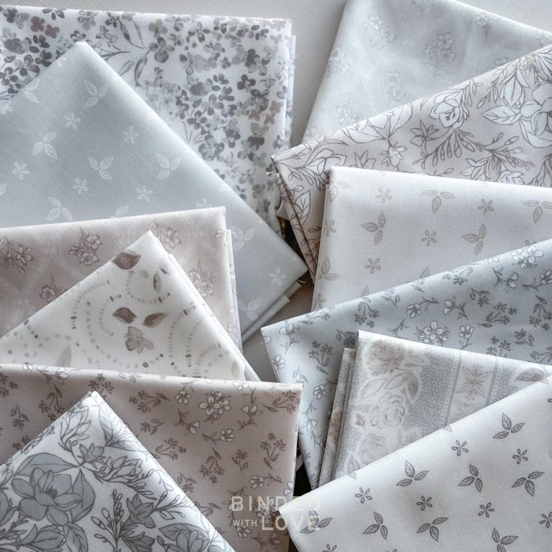Peut inclure: Un assortiment de morceaux de tissu pli&eacute;s dans des tons de gris et de blanc. Les tissus pr&eacute;sentent des motifs floraux et de feuilles, certains affichant de petits motifs de fleurs et de feuilles. La phrase "BIND WITH LOVE" est visible sur l'un des morceaux de tissu.