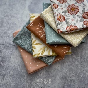 May include: A stack of folded fabric squares featuring various patterns including rust-colored flowers, white floral prints on sage green, a solid rust-colored cloth with white line accents, and a light pink fabric with white polka dots.