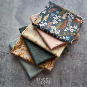 May include: A stack of six folded fabric squares featuring various patterns including a dark teal print with deer and trees, a solid pale pink, a cream fabric with small gold motifs, a dark green rose pattern, a mustard floral print, and solid light blue.