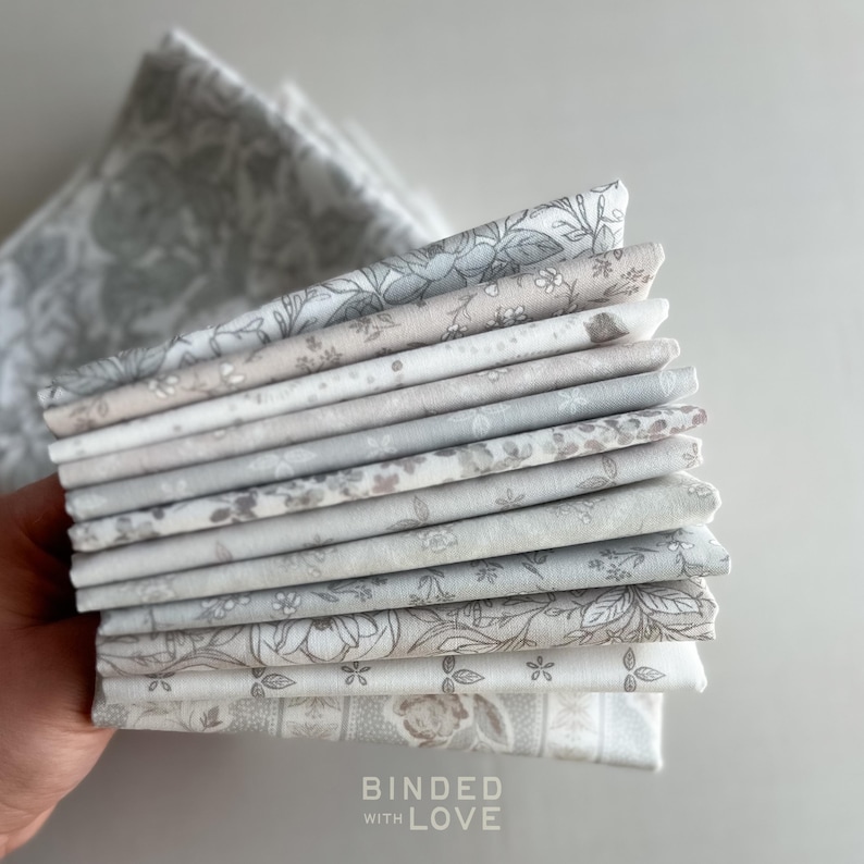 Peut inclure: Une pile de carr&eacute;s de tissu pli&eacute;s dans diverses nuances de gris et de blanc, avec des motifs floraux et botaniques. Les tissus sont tenus dans une main, avec les mots "BOUNDED WITH LOVE" visibles en bas.