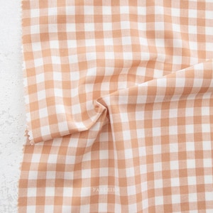 May include: Close-up of a fabric swatch with a classic gingham pattern. The fabric features a repeating pattern of squares in a warm peach and white color scheme. The fabric is slightly wrinkled, showing its texture.