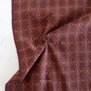 May include: Close-up of a folded fabric with a grid pattern. The fabric is a deep burgundy color with a repeating pattern of squares and rectangles in shades of brown and tan. The word "FABLEISM" is printed in white.