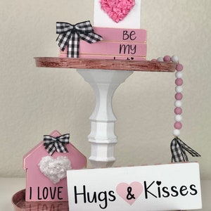 May include: A tiered tray decorated with pink and white wooden signs. The signs feature the phrases "Be my", "I love", and "Hugs & Kisses". The tray also includes a pink and white beaded garland and a pink house-shaped sign with a heart.