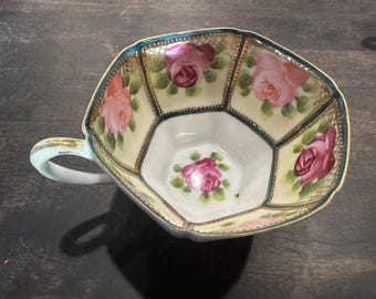 May include: An antique teacup with a unique geometric design. The cup features hand-painted pink roses on a cream-colored background, outlined with gold trim and a teal border. The handle is a light blue color.