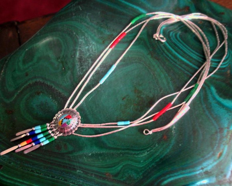 May include: A silver bolo tie with a central medallion featuring a colourful design. The tie has multiple strands with red, green, and blue accents, and silver tips. The bolo tie is displayed on a green, patterned surface.