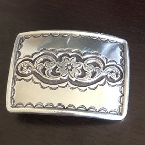 May include: Silver-toned rectangular belt buckle with intricate floral and scrollwork design. The buckle features a scalloped edge and a central flower motif, showcasing detailed craftsmanship. A classic accessory.