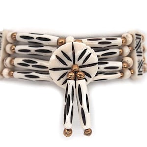 May include: Cream-colored bracelet with black oval patterns and gold-colored accents. The bracelet features a central circular design with radiating lines and dangling elements, showcasing a tribal or bohemian aesthetic.