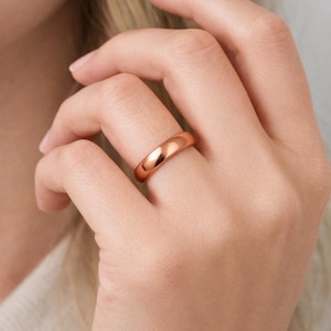 May include: A rose gold ring with a smooth, polished finish. The ring is a simple band design, likely for everyday wear. The ring is shown on a person's finger.