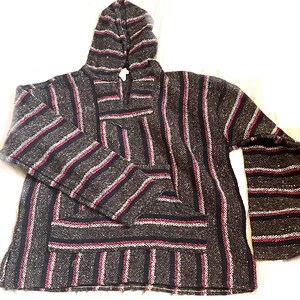 May include: A gray, hooded Baja hoodie with a front pocket. The striped pattern includes dark blue, white, and pink lines. The long-sleeved pullover is made of a woven fabric.