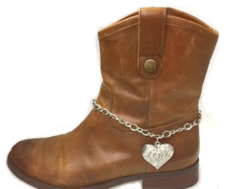 May include: A brown leather ankle boot with a silver chain and a heart-shaped charm. The boot has a round toe and a low heel. The charm is encrusted with small, clear stones.