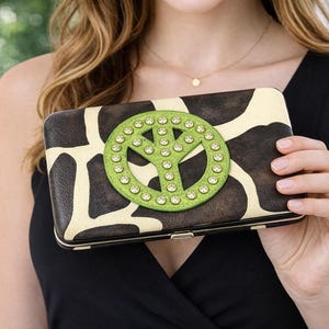 May include: A rectangular wallet with a giraffe print and a green peace sign embellished with rhinestones. The wallet has a silver frame and is held by a person wearing a black top.