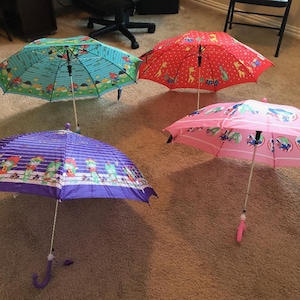 May include: Four open children's umbrellas in various colors: teal, red, pink, and purple. The teal umbrella has a cartoon design, the red one has a polka dot pattern, the pink one has a circular design, and the purple one has a repeating pattern.