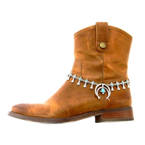May include: A brown leather ankle boot with a silver-toned chain ankle bracelet. The bracelet features turquoise-colored stones and decorative charms. The boot has a square toe and a small heel.