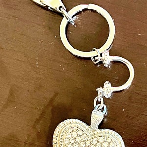 May include: Silver-toned keychain with a heart-shaped charm. The charm is encrusted with small, clear crystals and has a decorative border. The keychain includes a clasp and a split ring for attaching keys or other items.