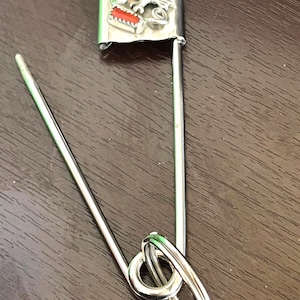 May include: A large silver safety pin with a decorative head featuring a turquoise stone, an eagle, and a red rectangular accent. The pin is open, displaying its long, curved needle and looped closure.