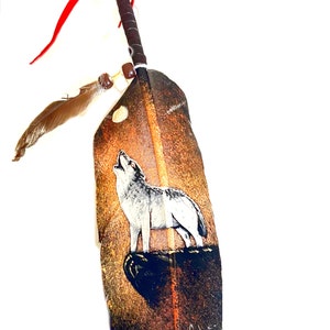 May include: A decorative feather with a painted image of a howling wolf. The feather is brown with a dark brown and black base. The handle is wrapped in dark brown leather with a red leather accent and a small feather attached.