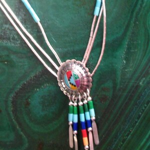 May include: A silver bolo tie necklace with a central, multi-coloured inlaid design. The pendant has a scalloped edge and hangs from three silver strands. The necklace features turquoise beads and colourful dangling accents.