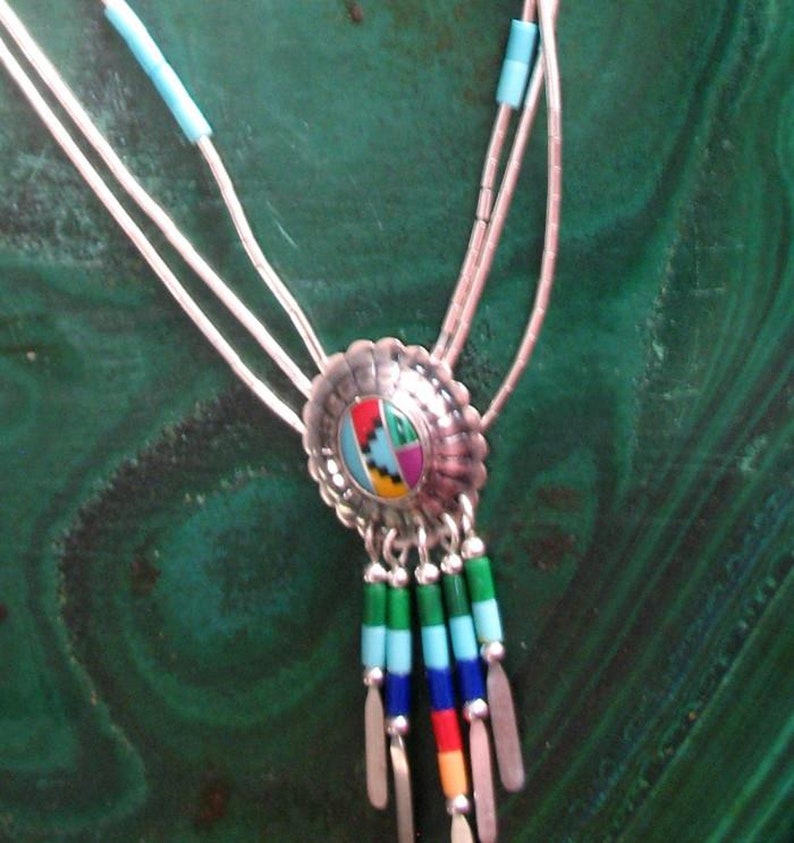 May include: A silver bolo tie necklace with a central, multi-coloured inlaid design. The pendant has a scalloped edge and hangs from three silver strands. The necklace features turquoise beads and colourful dangling accents.