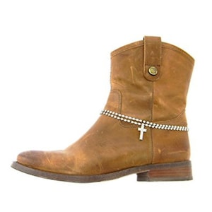 May include: A brown leather ankle boot with a decorative silver chain and a cross charm. The boot has a low heel and a rounded toe. The leather has a distressed look.