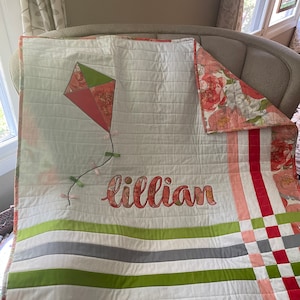 May include: A white baby quilt with green, red, and pink floral fabric. The quilt has a green, red, and pink kite design with a tail and the name "lillian" embroidered in pink.