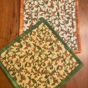 May include: Two quilted pot holders with a floral pattern. One pot holder has a green border and the other has an orange border.