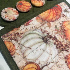 Può includere: Una pochette in tessuto verde con chiusura a zip e una fodera in tessuto a tema autunnale. La fodera presenta una zucca bianca, zucche arancioni e foglie autunnali. La pochette ha quattro bottoni sul davanti.