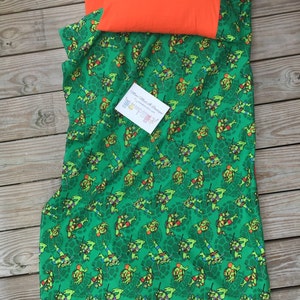 May include: A green and orange twin-sized sleeping bag with a Teenage Mutant Ninja Turtles print. The bag features a repeating pattern of the four turtles in action poses. The bag is made of soft, comfortable fabric and is perfect for sleepovers or camping trips.