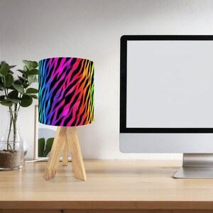 May include: A small table lamp with a vibrant, rainbow-coloured zebra print lampshade. The lampshade sits atop a light-coloured, three-legged wooden base. The lamp is on a wooden desk, next to a computer monitor and a vase with greenery.