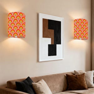 May include: Two wall sconces with a colorful rainbow pattern. The sconces are cylindrical and emit a warm light. A framed abstract art piece hangs between them, above a brown sofa with pillows.