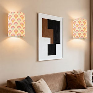 May include: Two wall sconces with a retro design, featuring a repeating pattern of pink, yellow, and peach arcs on a cream background. The sconces are mounted on a beige wall, with a framed abstract art piece and a brown sofa below.