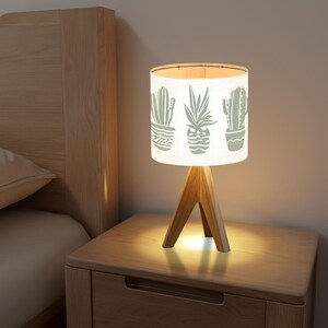 May include: A white table lamp with a wooden tripod base. The lampshade features a design of three potted cacti in a light gray color.