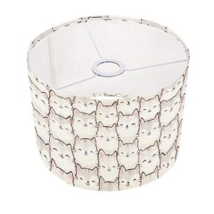 May include: A drum lampshade with a repeating pattern of smiling cat faces in grey and white. The lampshade has a white interior and a metal ring at the top for fitting to a lamp base. The design is playful and suitable for a child's room or a cat lover's home. The diameter is approximately 25 cm.