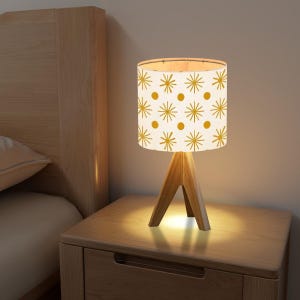 May include: A white table lamp with a wooden tripod base and a white lampshade featuring a geometric pattern of yellow stars and dots. The lamp is switched on and illuminating the bedside table.