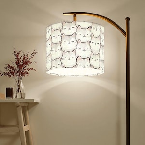 May include: A gold floor lamp with a white fabric lampshade featuring a repeating pattern of cartoon cat faces. The lampshade is cylindrical and has a white background with black outlines.