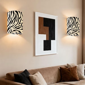 Könnte beinhalten: Zwei Wandleuchten mit Zebramuster sind an einer beigefarbenen Wand angebracht. Die Leuchten sind zylindrisch und strahlen ein warmes Licht aus. Zwischen ihnen hängt ein gerahmtes abstraktes Kunstwerk über einem braunen Sofa mit Kissen.