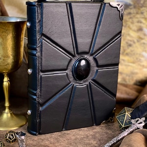 Puede incluir: Diario de cuero negro con un dise&ntilde;o geom&eacute;trico y una piedra negra central. El diario tiene detalles plateados en las esquinas y un lomo azul oscuro con remaches plateados. Un c&aacute;liz dorado y dados en el fondo.