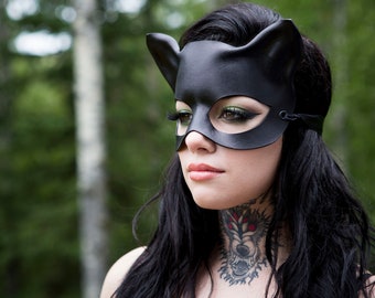 May include: A woman wearing a black leather cat mask with ears. She has dark hair and a wolf tattoo on her neck.
