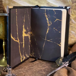 Puede incluir: Cuaderno negro abierto con vetas doradas en las p&aacute;ginas interiores. El cuaderno tiene una cinta negra como marcador y protectores de esquina met&aacute;licos. Un bol&iacute;grafo plateado, una llave y dados est&aacute;n sobre la superficie marr&oacute;n.